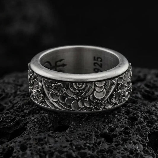 An oxidized sterling silver spring flower spinner ring rests on dark volcanic rock, showing raised floral blossoms, curved petal patterns, and dark recessed texture in a macro studio close-up.