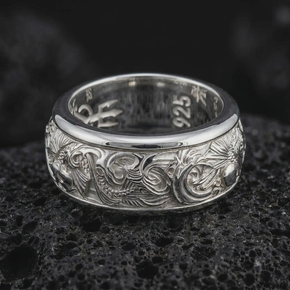 The polished silver carp and wave wedding band is displayed on textured black volcanic rock, highlighting the sculpted fish motifs, ornamental water lines, and wide spinner ring profile in a premium macro photograph.