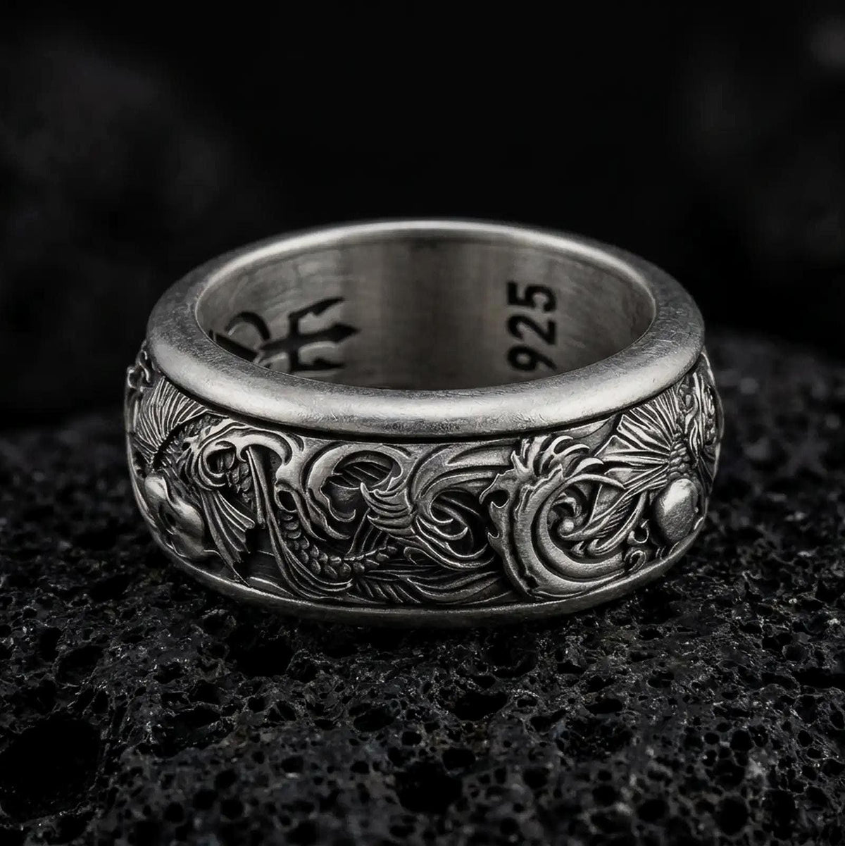 An oxidized sterling silver koi fish spinner ring rests on dark volcanic rock, showing raised carp details, flowing wave patterns, and a dark recessed band background in a macro studio close-up.