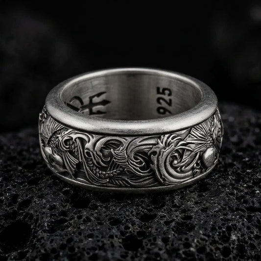An oxidized sterling silver koi fish spinner ring rests on dark volcanic rock, showing raised carp details, flowing wave patterns, and a dark recessed band background in a macro studio close-up.