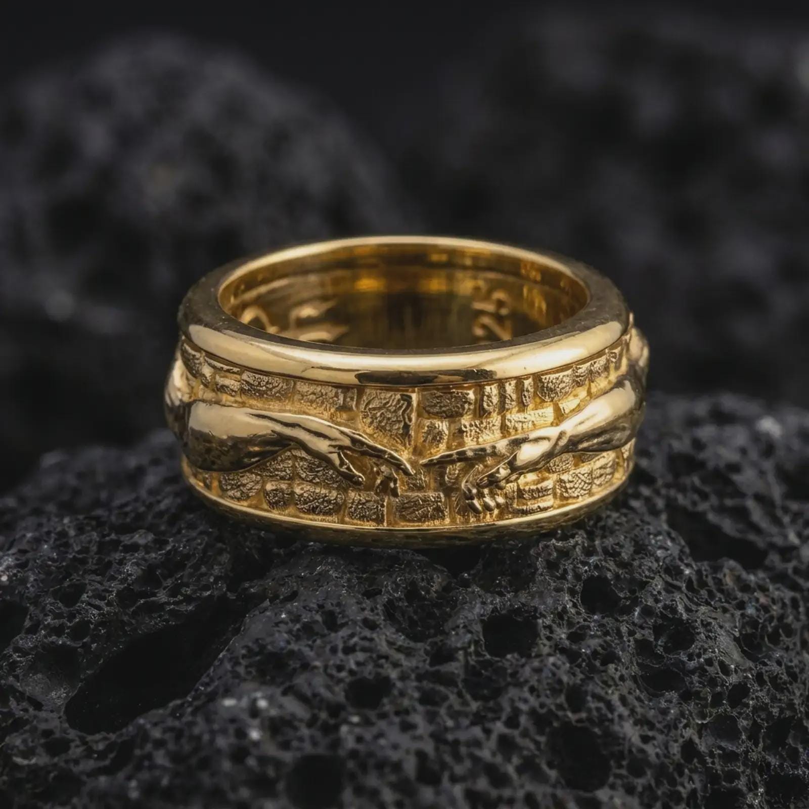 A gold plated reaching hands spinner ring rests on rugged volcanic rock, featuring the Creation of Adam inspired design, dimensional hand relief, and wide wedding band structure in a luxury macro close-up.