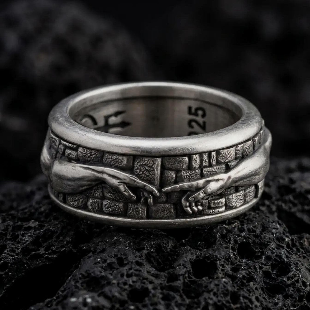 An oxidized sterling silver Creation of Adam spinner ring rests on dark volcanic rock, showing sculpted reaching hands, textured wall details, and a wide symbolic band in a macro studio close-up.