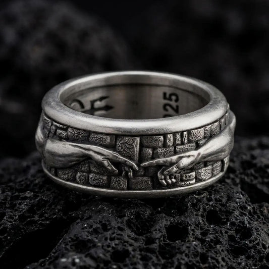An oxidized sterling silver Creation of Adam spinner ring rests on dark volcanic rock, showing sculpted reaching hands, textured wall details, and a wide symbolic band in a macro studio close-up.