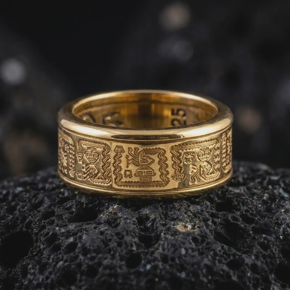 A gold plated Aztec spinner band rests on rugged volcanic rock, featuring detailed geometric pattern panels and an engravable wide ring structure in a luxury macro studio close-up.