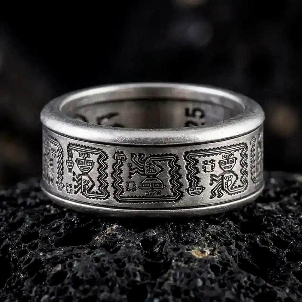 An oxidized sterling silver Aztec spinner ring rests on dark volcanic rock, showing engraved geometric panels, dark recessed details, and a wide symbolic band in a macro studio close-up.