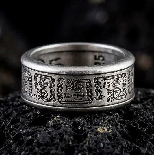 An oxidized sterling silver Aztec spinner ring rests on dark volcanic rock, showing engraved geometric panels, dark recessed details, and a wide symbolic band in a macro studio close-up.