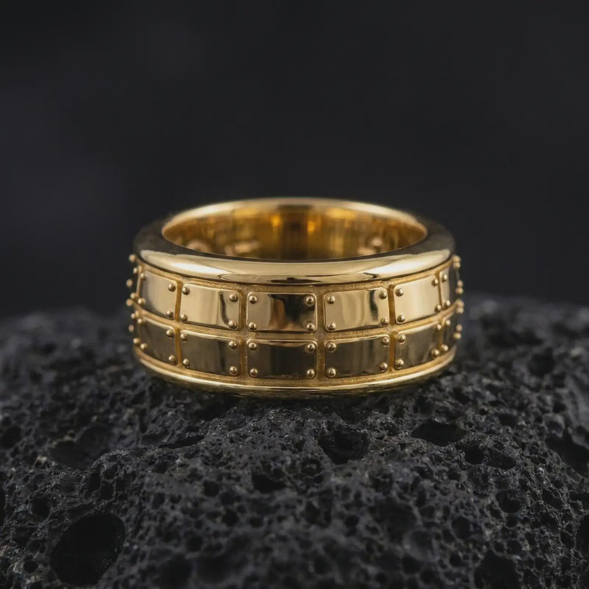 A gold plated geometric spinner ring sits on textured volcanic rock, showcasing the bold plate motif, structured fidget band design, and luxurious metallic finish in a premium macro studio image.