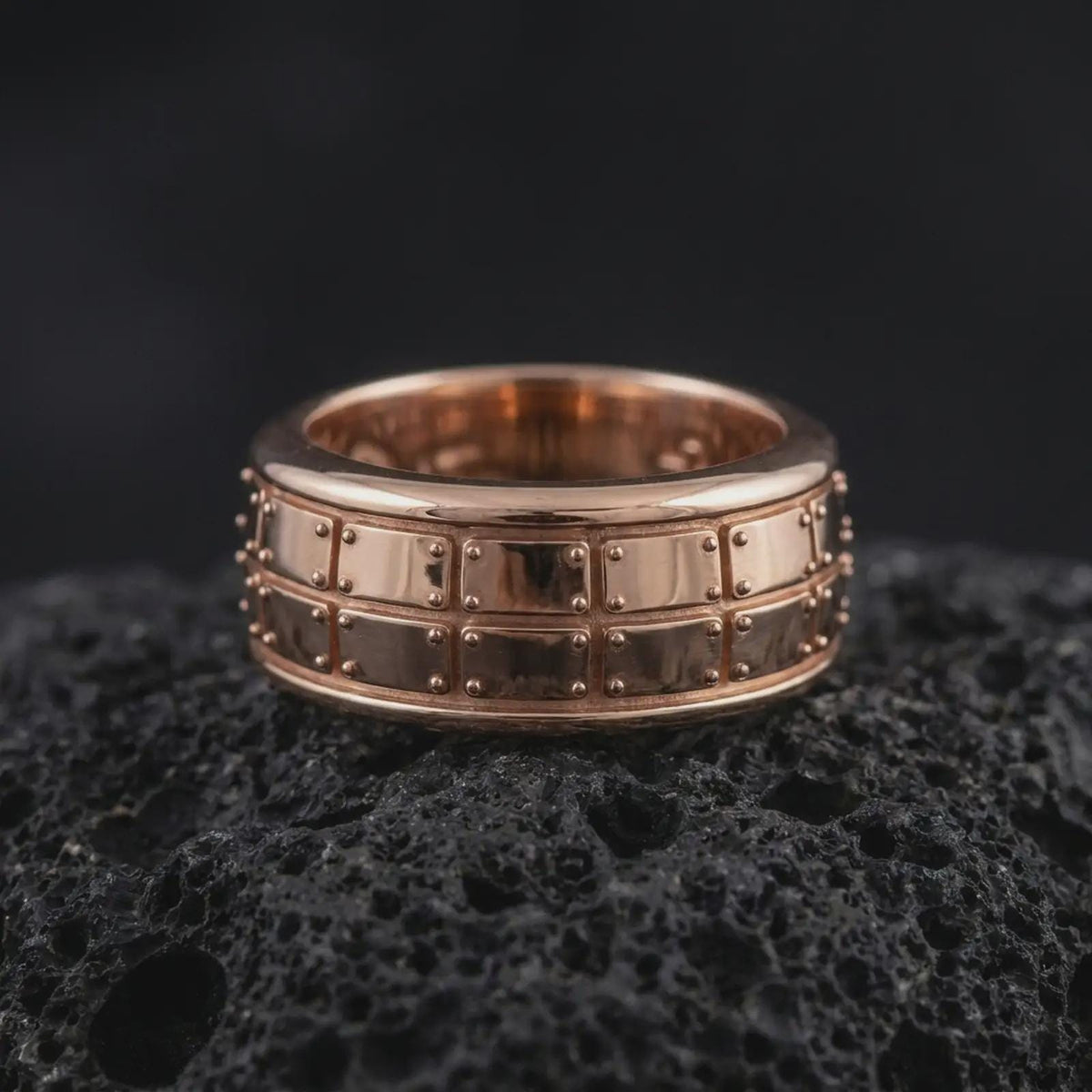 A rose gold plated plate pattern spinner ring is arranged on dark volcanic rock, featuring the segmented geometric surface, rotating band profile, and warm elegant finish in a close-up jewelry photograph.