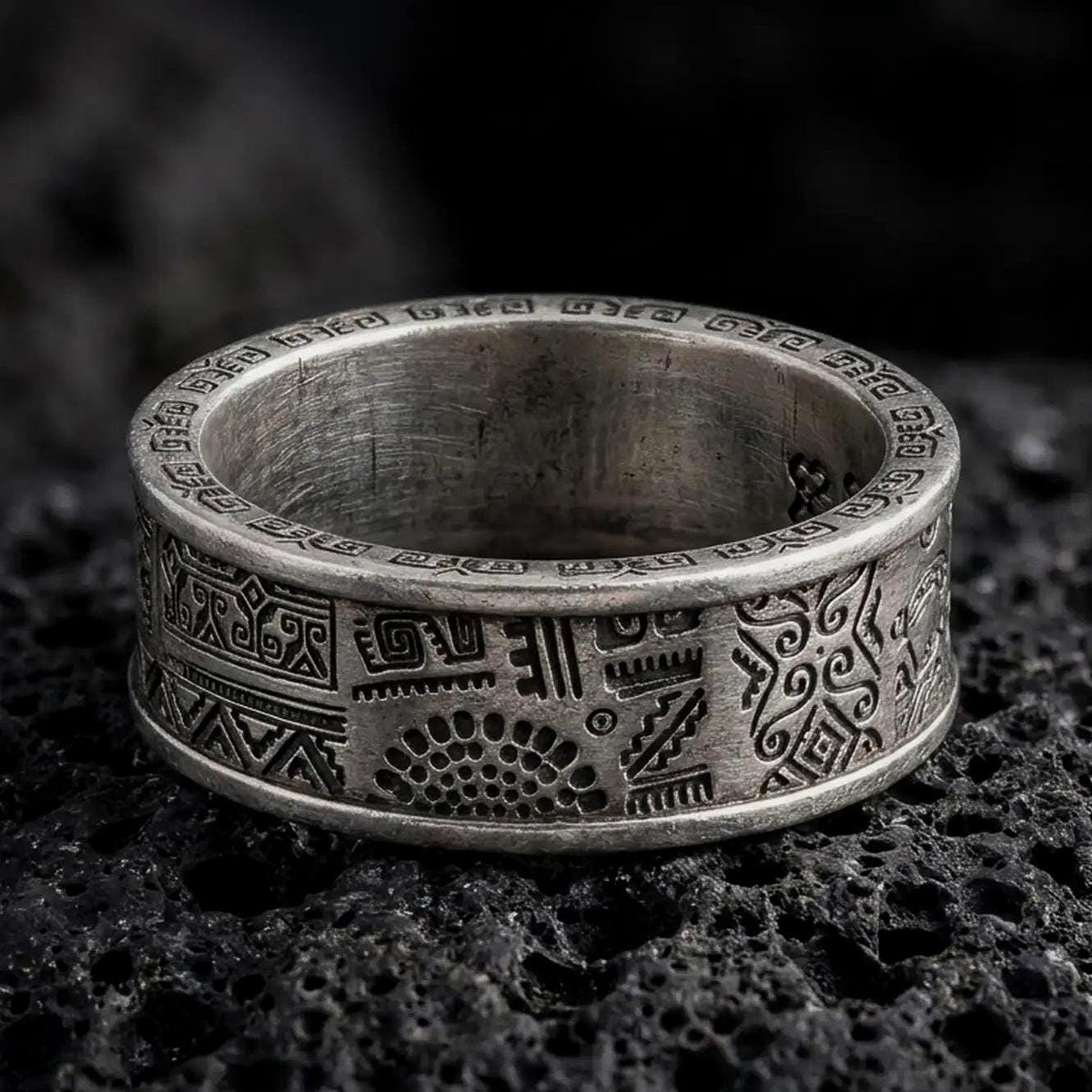 An oxidized sterling silver Aztec spinner ring rests on dark volcanic rock, showing geometric panels, stepped motifs, tribal-inspired symbols, and dark recessed engravings in a macro studio close-up.