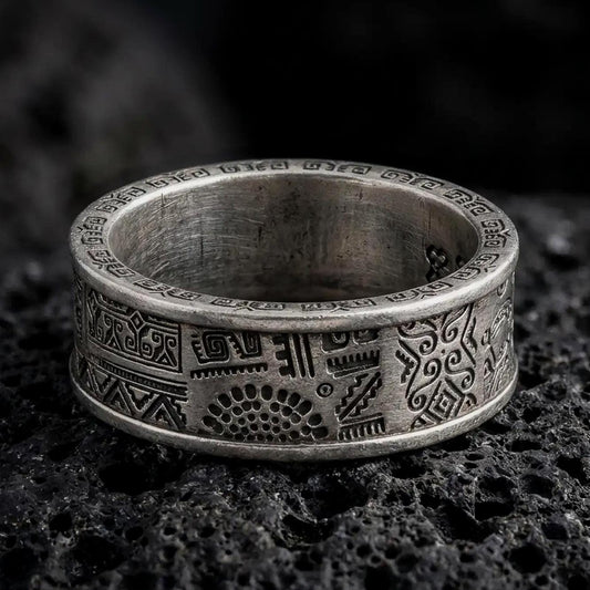 An oxidized sterling silver Aztec spinner ring rests on dark volcanic rock, showing geometric panels, stepped motifs, tribal-inspired symbols, and dark recessed engravings in a macro studio close-up.