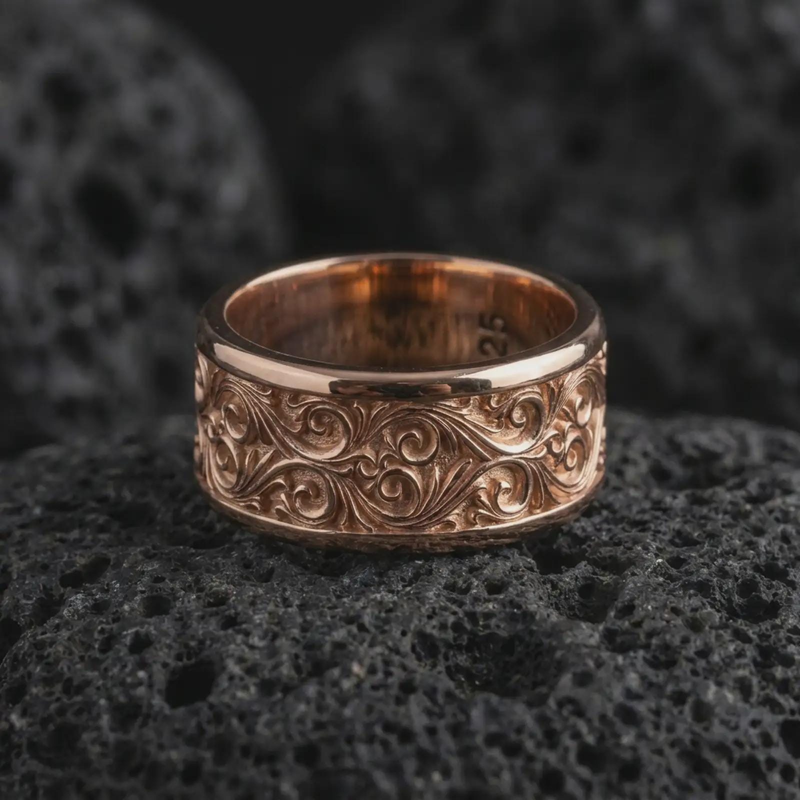 The rose gold plated scrollwork wedding band is arranged on black volcanic rock, emphasizing its flowing ornamental relief, vintage-inspired texture, and refined unisex spinner design in a detailed macro image.