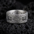 Load image into Gallery viewer, An oxidized sterling silver ornate cross band ring rests on dark volcanic rock, featuring raised cross motifs, intricate scrollwork, and a detailed floral centerpiece in a macro studio close-up.
