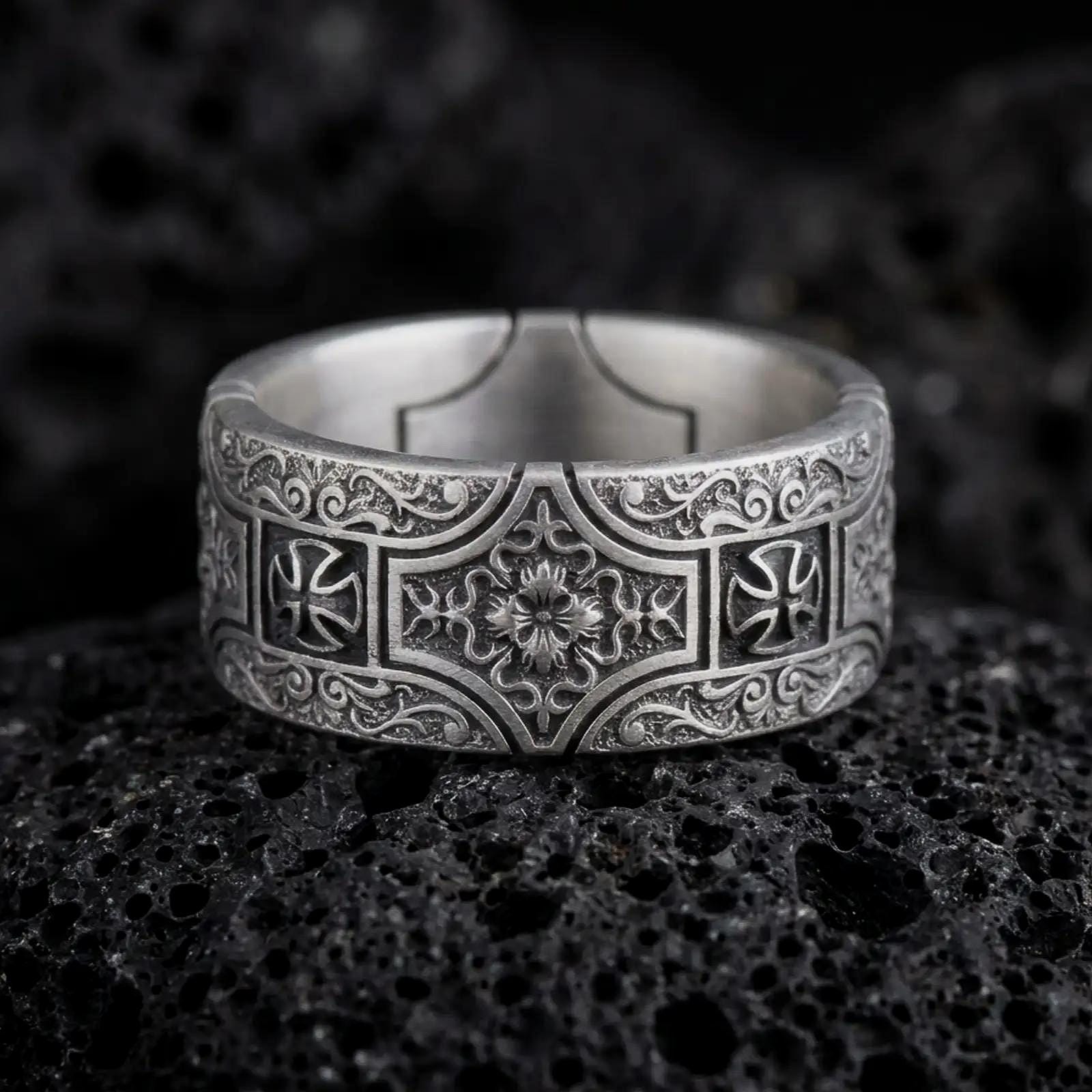 An oxidized sterling silver ornate cross band ring rests on dark volcanic rock, featuring raised cross motifs, intricate scrollwork, and a detailed floral centerpiece in a macro studio close-up.