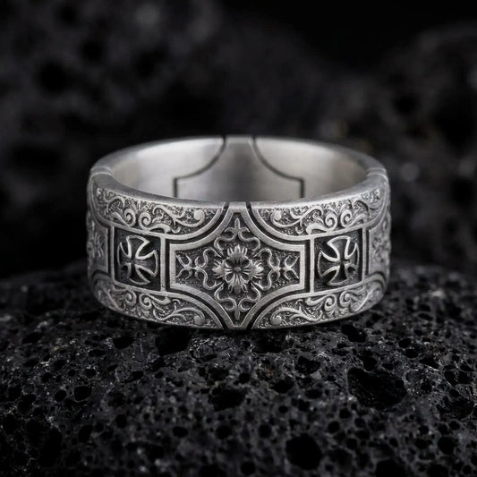 An oxidized sterling silver ornate cross band ring rests on dark volcanic rock, featuring raised cross motifs, intricate scrollwork, and a detailed floral centerpiece in a macro studio close-up.