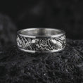 Load image into Gallery viewer, The polished silver botanical wedding band is displayed on textured black volcanic rock, highlighting its raised floral relief, flowing leaves, smooth rounded edges, and continuous nature-inspired pattern in a premium macro photograph.
