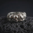 Load image into Gallery viewer, The polished silver skull and rose ring is displayed on textured black volcanic rock, highlighting the three-dimensional skull relief, floral detailing, smooth band edges, and intricate gothic craftsmanship in a premium macro photograph.
