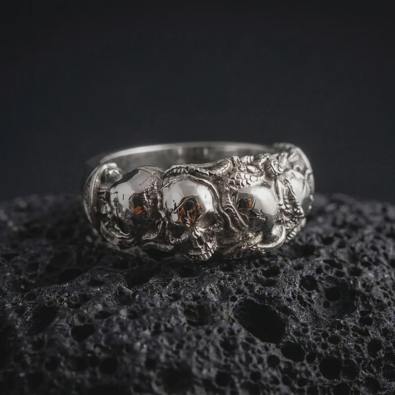 The polished silver skull and rose ring is displayed on textured black volcanic rock, highlighting the three-dimensional skull relief, floral detailing, smooth band edges, and intricate gothic craftsmanship in a premium macro photograph.