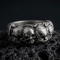 Load image into Gallery viewer, An oxidized sterling silver skull ring rests on dark volcanic rock, featuring a detailed raised skull surrounded by sculpted roses, leaves, and gothic floral elements in a dramatic macro studio close-up.
