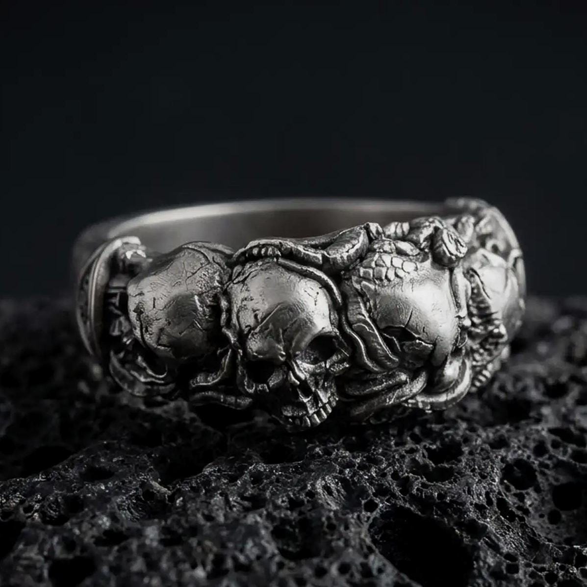 An oxidized sterling silver skull ring rests on dark volcanic rock, featuring a detailed raised skull surrounded by sculpted roses, leaves, and gothic floral elements in a dramatic macro studio close-up.