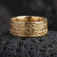 Load image into Gallery viewer, A gold plated Elven Star wedding band rests on rugged volcanic rock, showcasing the central geometric star, flowing fantasy knotwork, and dimensional wide band design in a luxury macro studio close-up.
