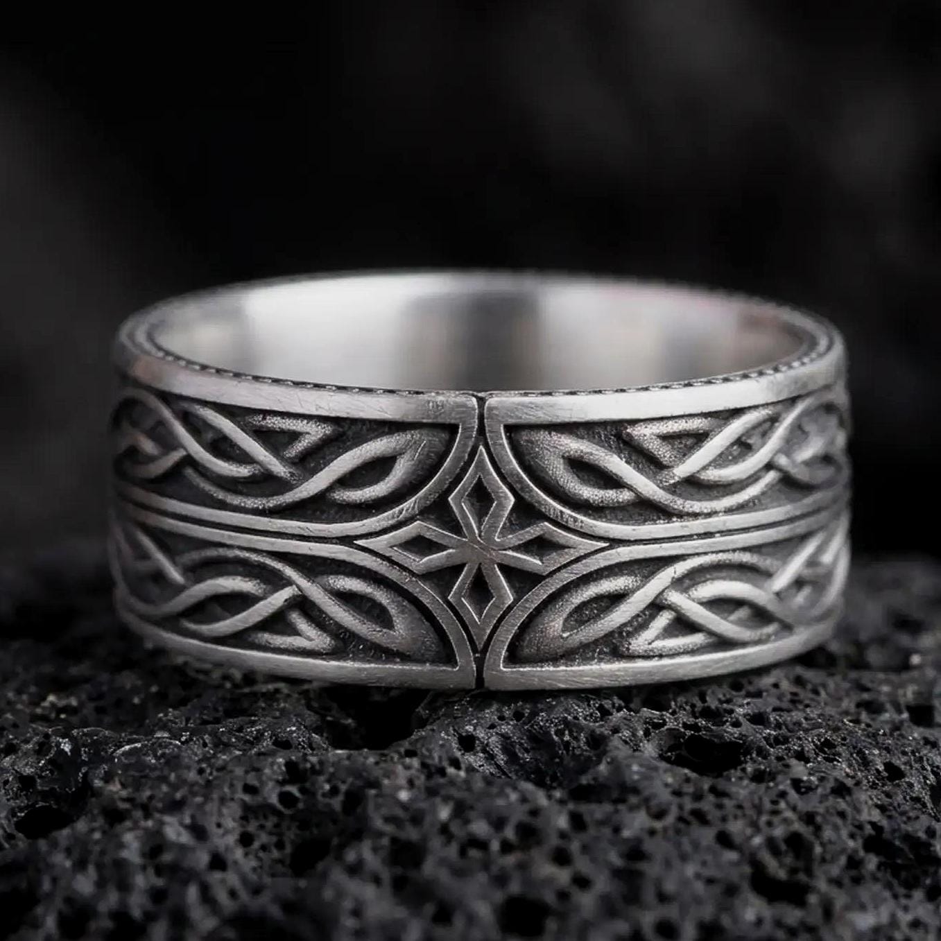 An oxidized sterling silver Elven Star ring rests on dark volcanic rock, featuring a geometric central star, flowing interwoven knotwork, and a dark recessed wide band in a macro studio close-up.