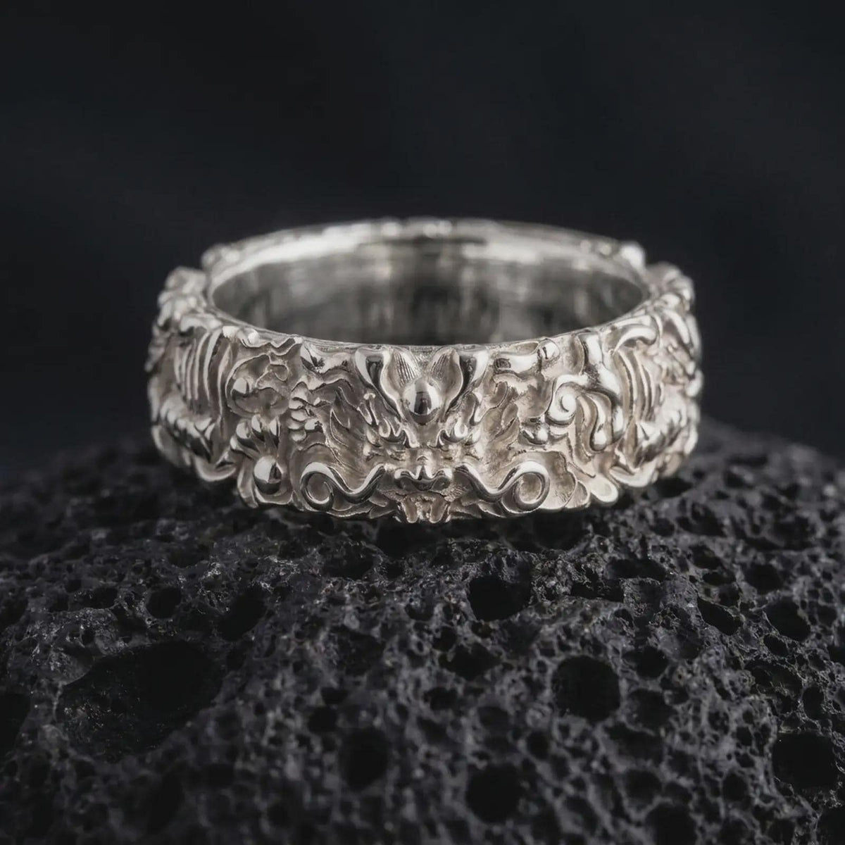 The polished silver mythical dragon ring is displayed on textured black volcanic rock, highlighting the raised dragon face, flowing ornamental details, sculpted scales, and substantial wide band profile in a premium macro photograph.
