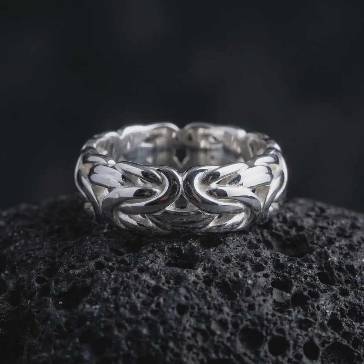 The polished silver Byzantine chain ring is displayed on textured black volcanic rock, highlighting the dimensional woven links, smooth interior, and bold handcrafted band structure in a premium macro photograph.
