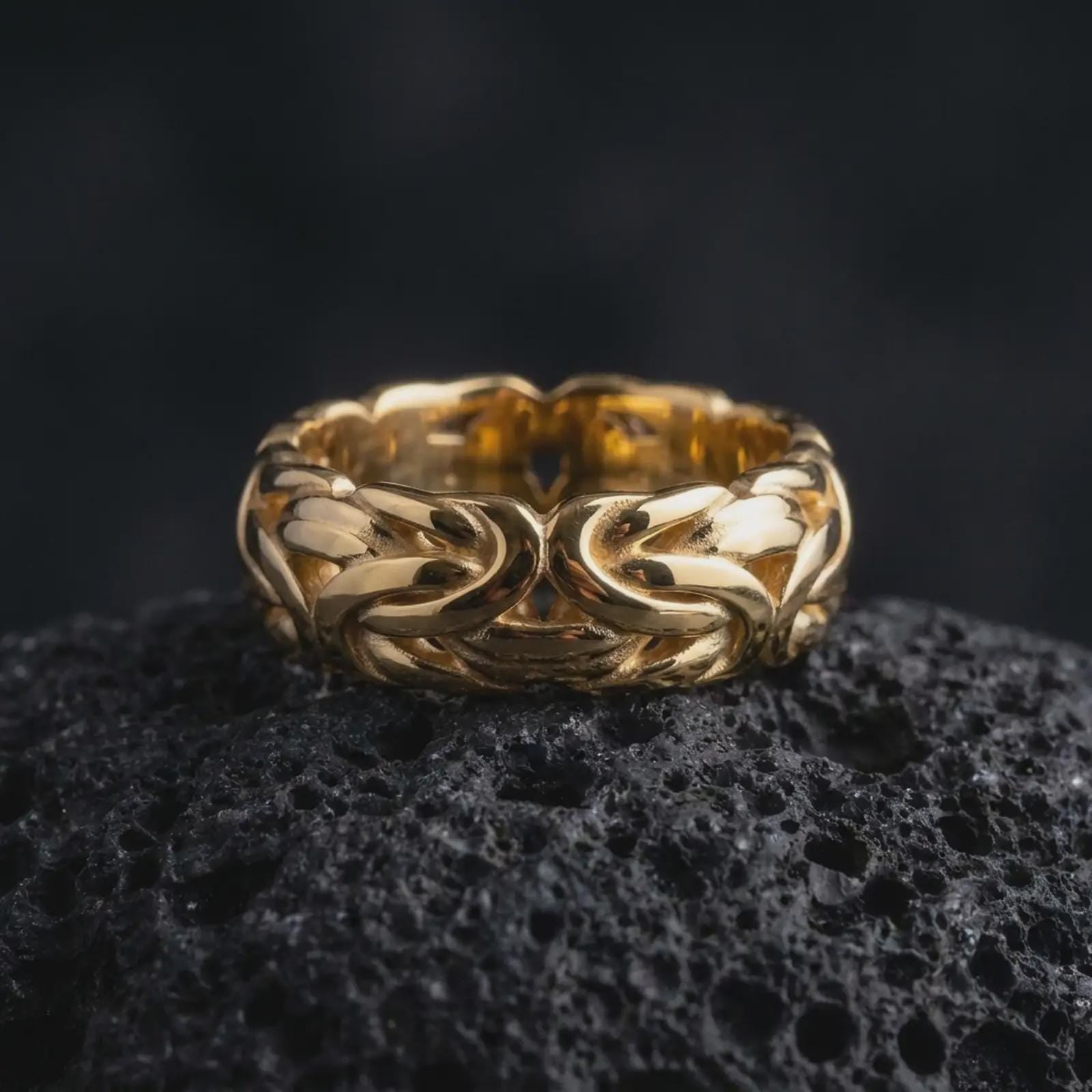 A gold plated Byzantine chain band ring rests on rugged volcanic rock, featuring interwoven chain-inspired links, strong sculptural depth, and a substantial unisex ring profile in a luxury macro studio close-up.