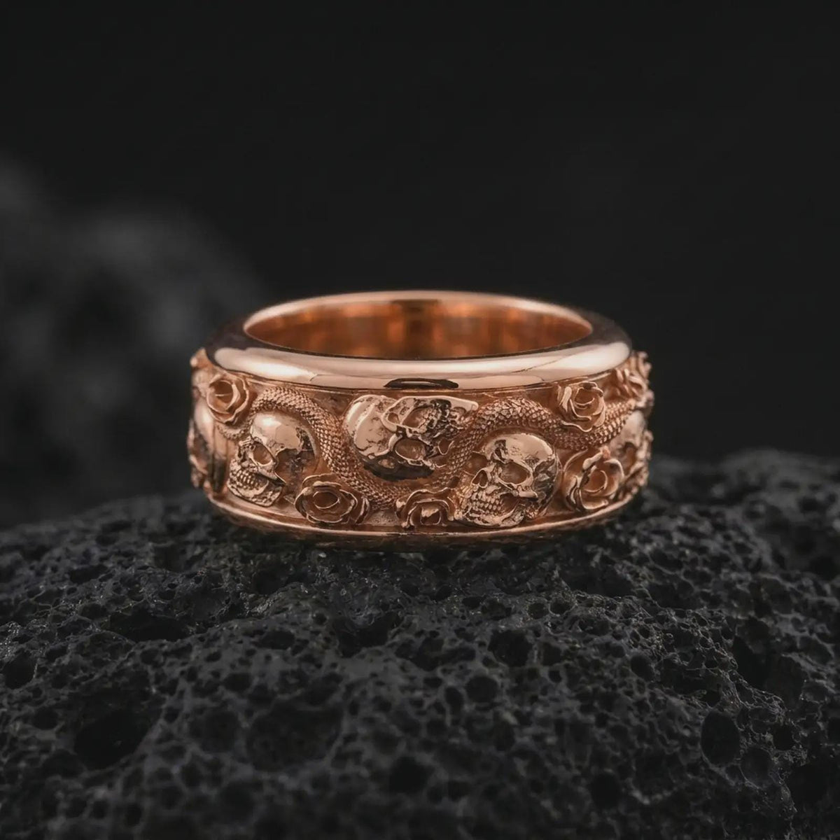 The rose gold plated skull and serpent band is arranged on black volcanic rock, emphasizing its sculpted skulls, winding snake, floral rose details, and bold dark romantic jewelry design in a detailed macro image.