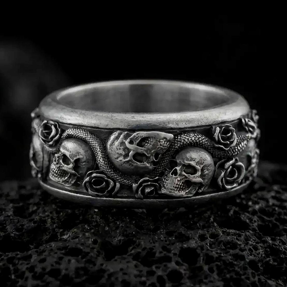 An oxidized sterling silver skull snake rose ring rests on dark volcanic rock, featuring sculpted skulls, a winding serpent, raised roses, and deep recessed detailing in a dramatic macro studio close-up.