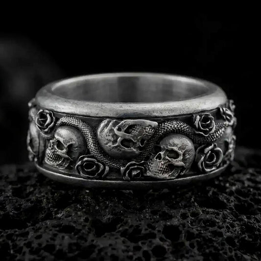 An oxidized sterling silver skull snake rose ring rests on dark volcanic rock, featuring sculpted skulls, a winding serpent, raised roses, and deep recessed detailing in a dramatic macro studio close-up.