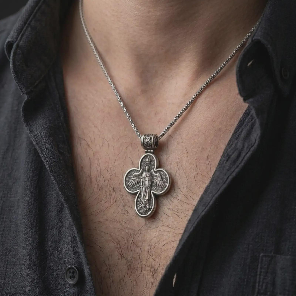 An oxidized sterling silver Saint Michael cross pendant is worn on the neck, displaying the double sided Christian design, protective symbolism, and detailed sculptural cross form in a close-up lifestyle photograph.