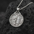 Load image into Gallery viewer, The polished silver crusader knight pendant is displayed on textured black volcanic rock, highlighting the raised armor, sword, cross-bearing shield, medieval architecture, and detailed medallion construction in a premium macro photograph.
