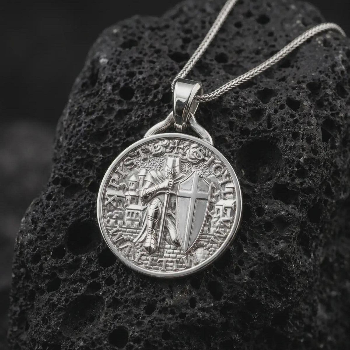 The polished silver crusader knight pendant is displayed on textured black volcanic rock, highlighting the raised armor, sword, cross-bearing shield, medieval architecture, and detailed medallion construction in a premium macro photograph.