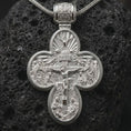 Load image into Gallery viewer, The polished silver Orthodox cross pendant is displayed on textured black volcanic rock, highlighting the double sided religious design, raised sculptural details, and refined sterling silver finish in a premium macro photograph.
