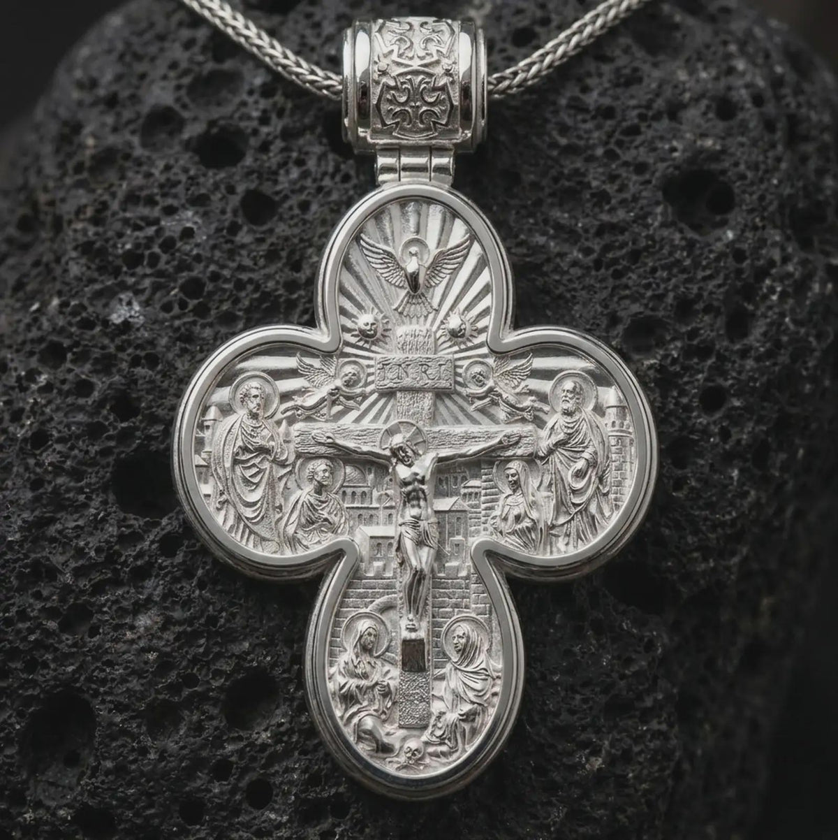 The polished silver Orthodox cross pendant is displayed on textured black volcanic rock, highlighting the double sided religious design, raised sculptural details, and refined sterling silver finish in a premium macro photograph.