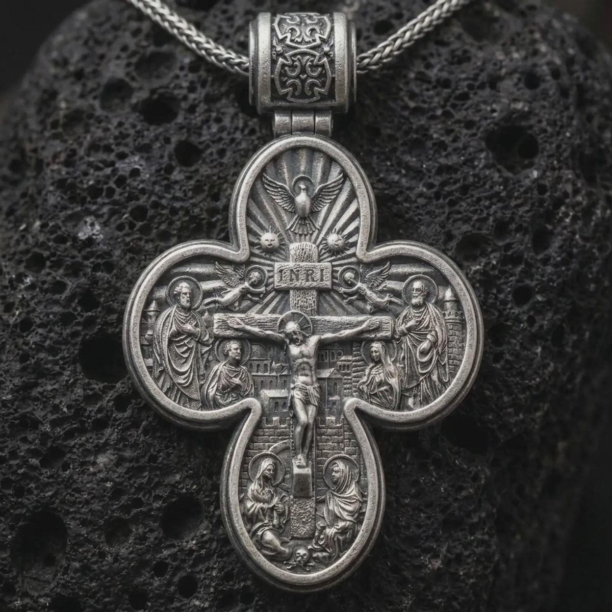 An oxidized sterling silver double sided Saint Michael cross pendant rests on dark volcanic rock, showing a detailed crucifixion scene with sacred figures, radiant motifs, and bold relief in a dramatic macro studio close-up.