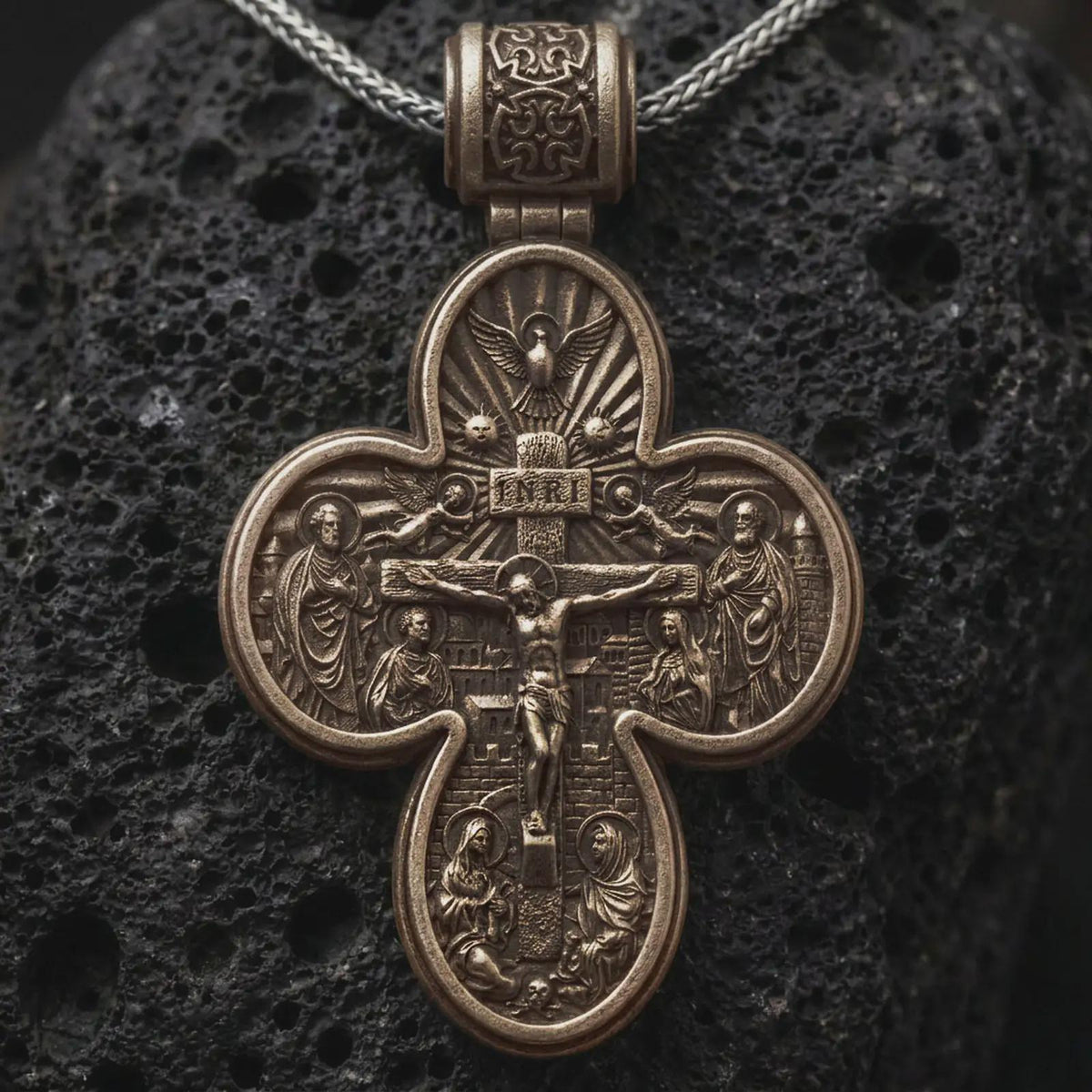 Bronze sterling silver double sided Saint Michael cross pendant rests on dark volcanic rock, showing a detailed crucifixion scene with sacred figures, radiant motifs, and bold relief in a dramatic macro studio close-up.