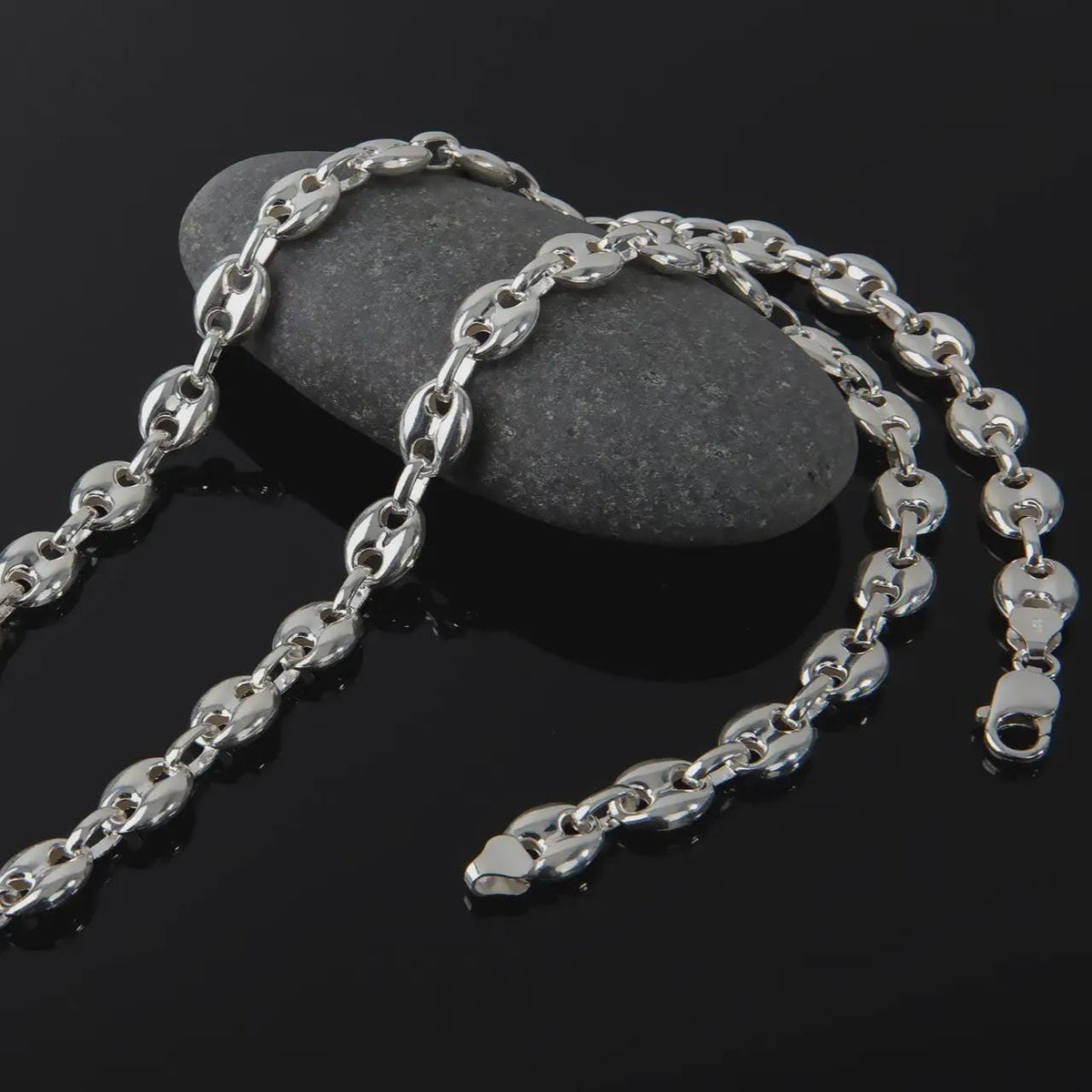 The polished silver 11mm Mariner chain necklace is displayed on textured black volcanic rock, highlighting the smooth puffed anchor links, reflective sterling silver surfaces, and bold wide-link construction in a premium macro photograph.
