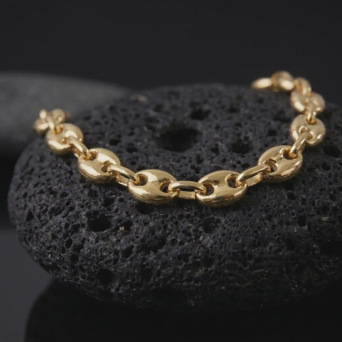 A gold plated 11mm Mariner chain bracelet rests on rugged volcanic rock, showcasing broad oval links, distinctive center bars, and a substantial nautical-inspired bracelet design in a luxury macro studio close-up.