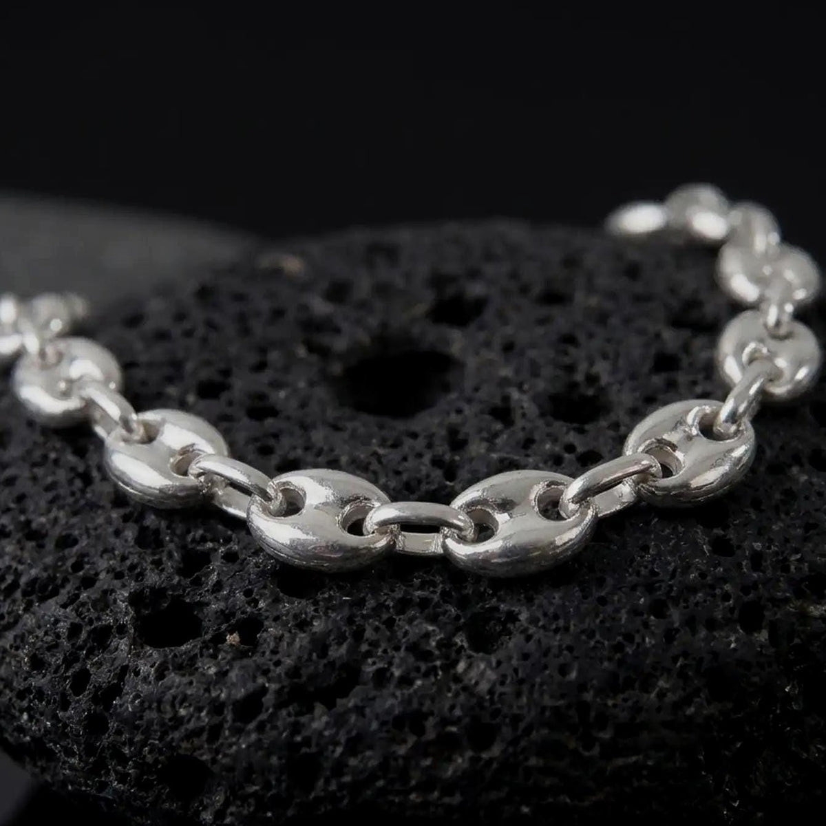 The polished silver 11mm Mariner bracelet is displayed on textured black volcanic rock, highlighting the smooth puffed anchor links, reflective surfaces, and bold wide-link construction in a premium macro photograph.