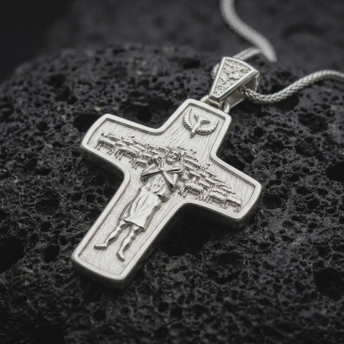 The polished silver Good Shepherd cross pendant is displayed on textured black volcanic rock, highlighting the shepherd figure, flock of sheep, dove motif, raised relief, and smooth sterling silver border in a premium macro photograph.
