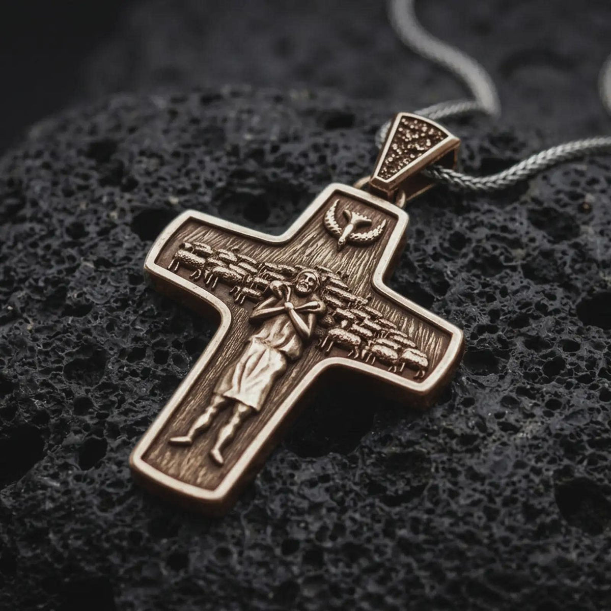 A Bronze sterling silver Pope Francis cross pendant rests on dark volcanic rock, featuring the Good Shepherd carrying a sheep, surrounding flock, dove symbol, and detailed recessed Christian imagery in a macro studio close-up.