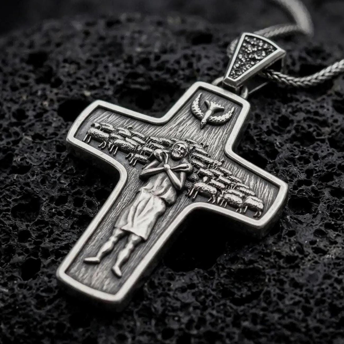 An oxidized sterling silver Pope Francis cross pendant rests on dark volcanic rock, featuring the Good Shepherd carrying a sheep, surrounding flock, dove symbol, and detailed recessed Christian imagery in a macro studio close-up.