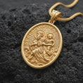 Load image into Gallery viewer, A gold plated St. Christopher pendant rests on rugged volcanic rock, showcasing the patron saint of travelers carrying the Christ Child, wave details, and symbolic Catholic protection imagery in a luxury macro studio close-up.
