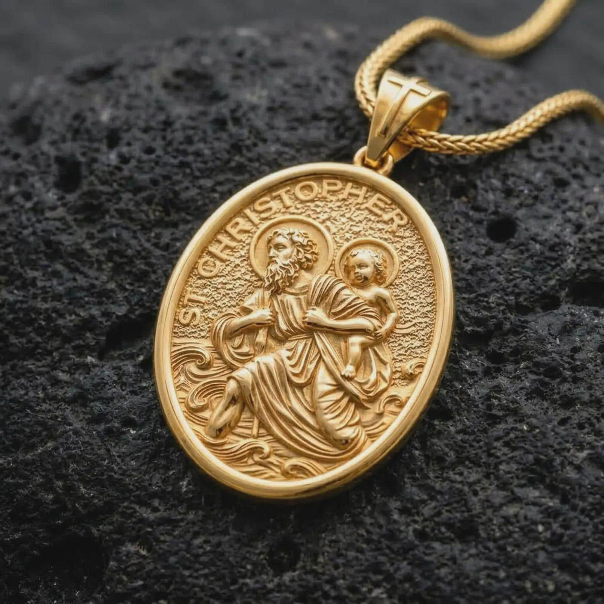 A gold plated St. Christopher pendant rests on rugged volcanic rock, showcasing the patron saint of travelers carrying the Christ Child, wave details, and symbolic Catholic protection imagery in a luxury macro studio close-up.