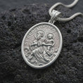 Load image into Gallery viewer, The polished silver St. Christopher medal is displayed on textured black volcanic rock, highlighting the haloed saint, Christ Child, flowing waves, oval medallion border, and detailed devotional relief in a premium macro photograph.
