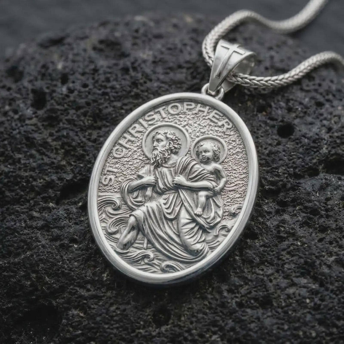 The polished silver St. Christopher medal is displayed on textured black volcanic rock, highlighting the haloed saint, Christ Child, flowing waves, oval medallion border, and detailed devotional relief in a premium macro photograph.