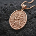 Load image into Gallery viewer, The rose gold plated St. Christopher necklace is arranged on black volcanic rock, emphasizing the sculpted saint and child figures, halo details, flowing water, and meaningful travelers protection design in a refined macro image.
