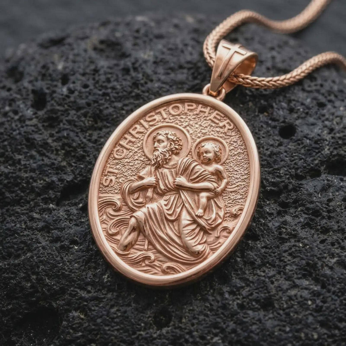 The rose gold plated St. Christopher necklace is arranged on black volcanic rock, emphasizing the sculpted saint and child figures, halo details, flowing water, and meaningful travelers protection design in a refined macro image.